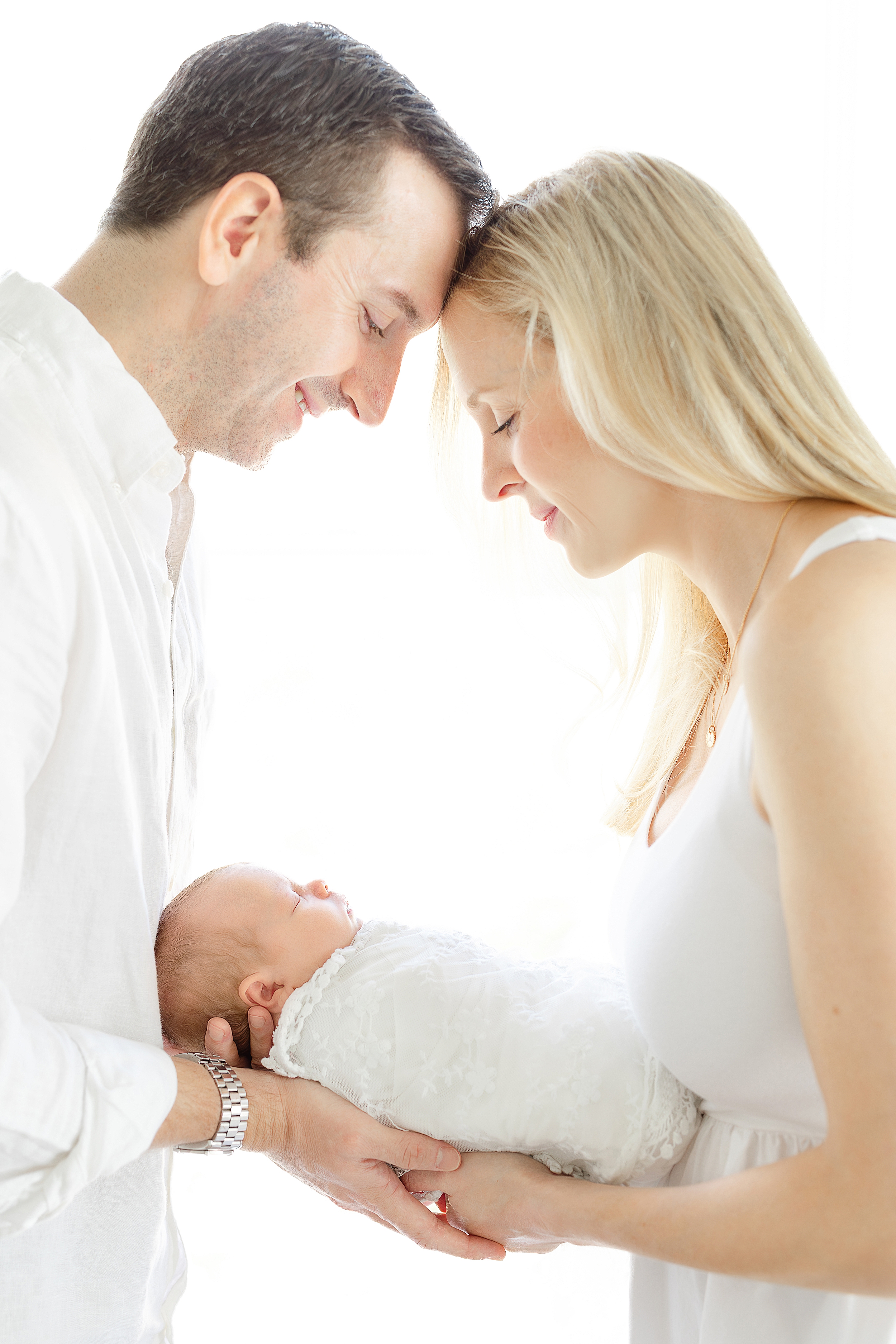 A man and a woman dressed in white cradle their newborn baby girl in front of a white lit window.