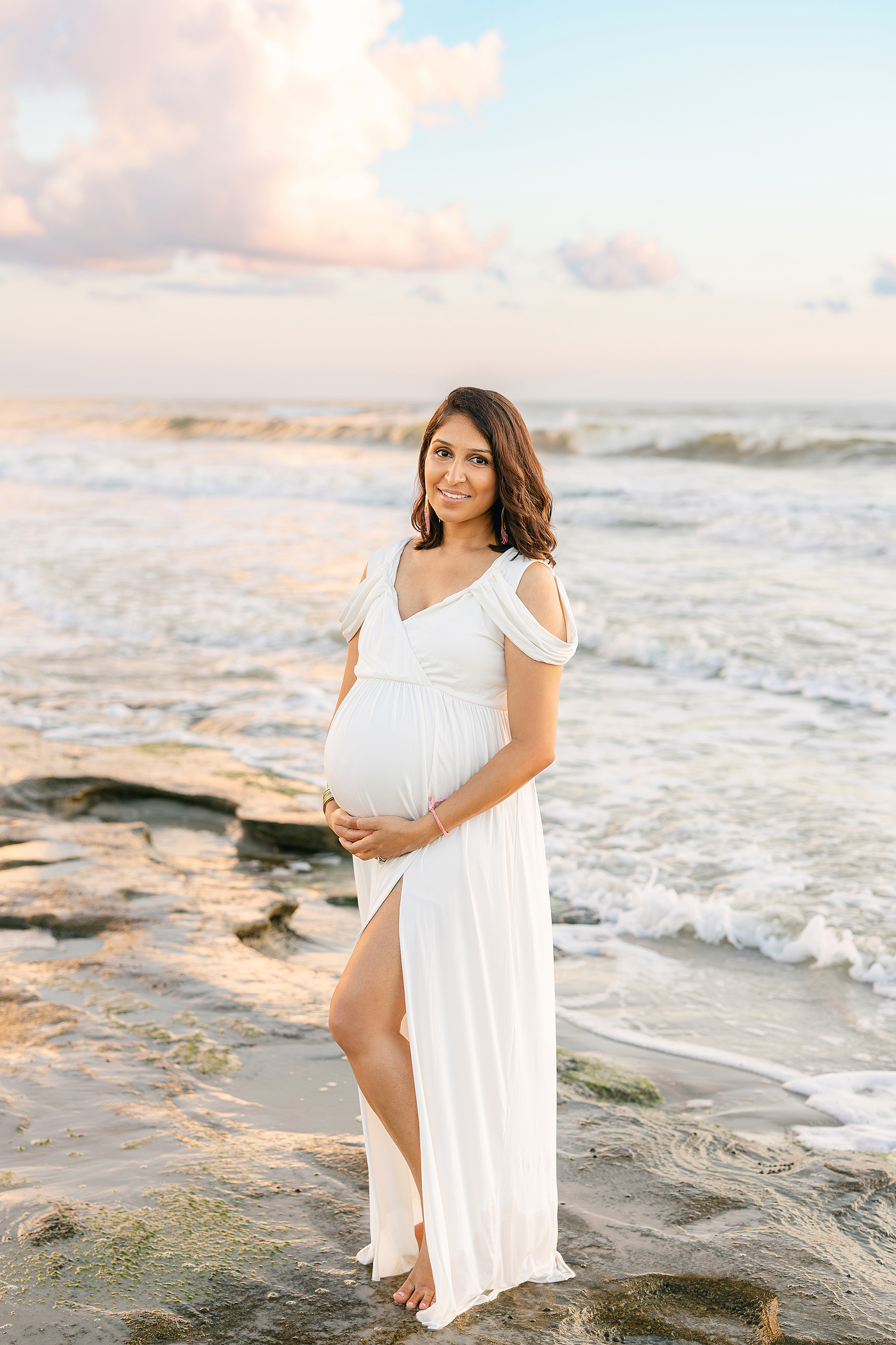 A colorful, sunrise maternity photo of a woman in a long white dress on the beach.