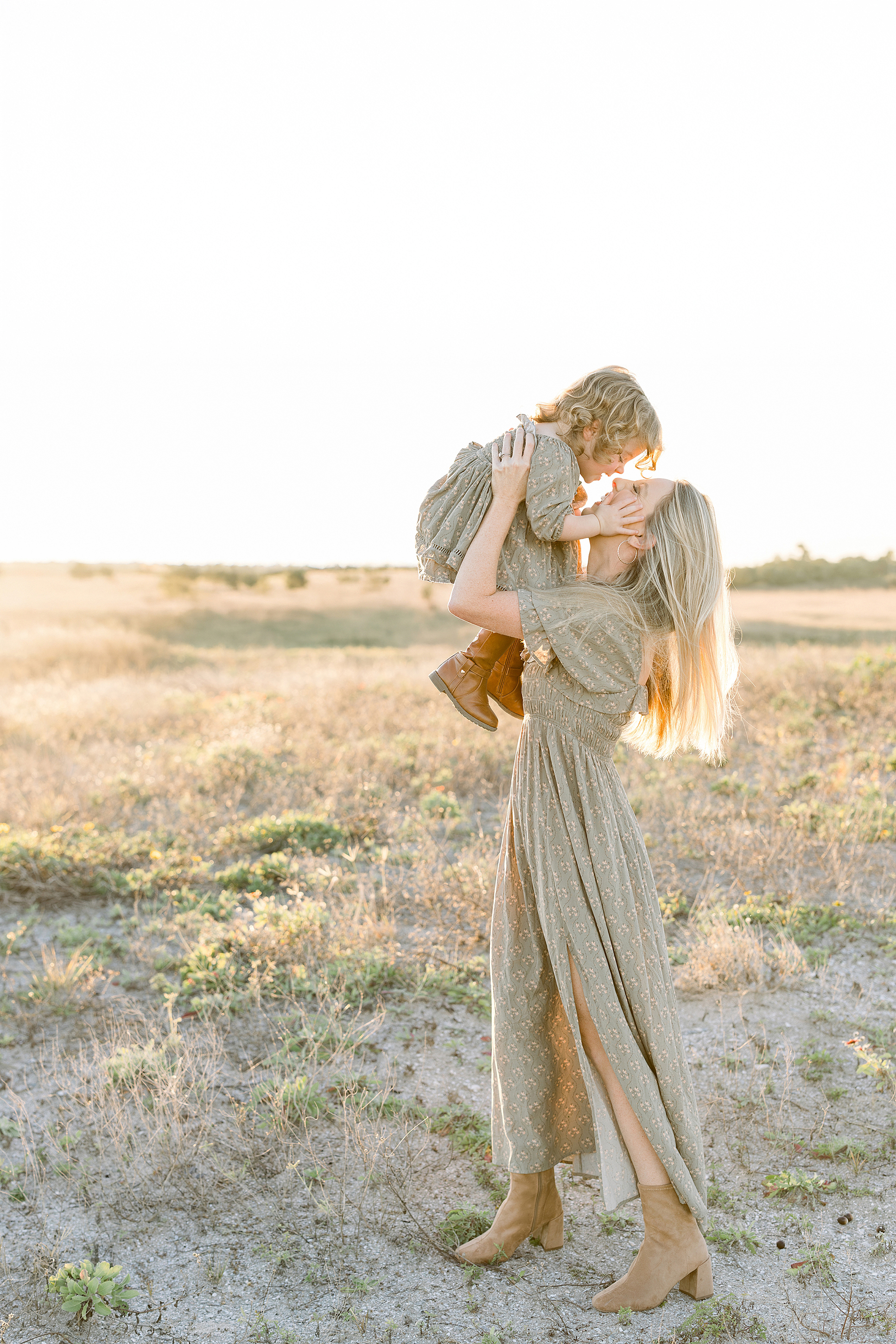 A woman in a green maxi dress holds up her toddler daughter also wearing green.