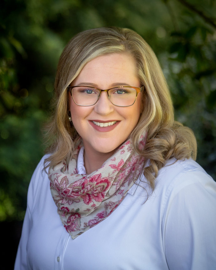 Smiling woman wearing glasses and a floral scarf, standing outdoors with a blurred green background.