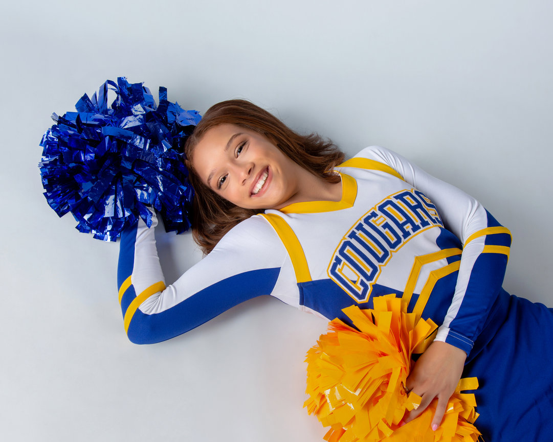 Cheerleader in blue and yellow uniform holding blue and yellow pom-poms, smiling while lying on the floor.
