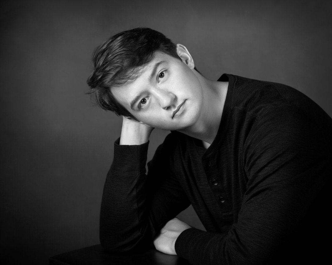 Young person in a dark shirt, leaning on a table, looking at camera, against a plain background in black and white.