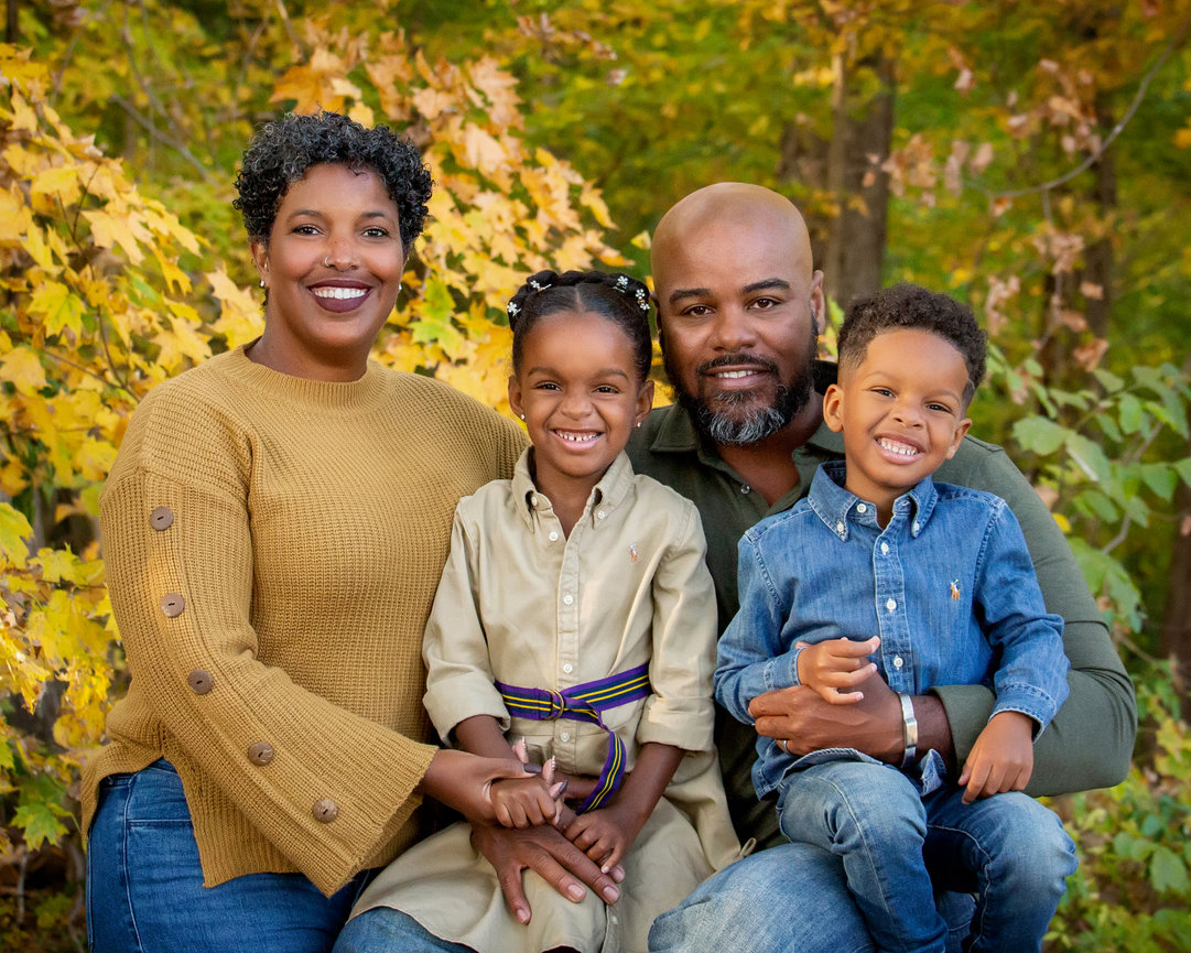 Family of four smiling in a park during autumn, surrounded by colorful fall foliage.