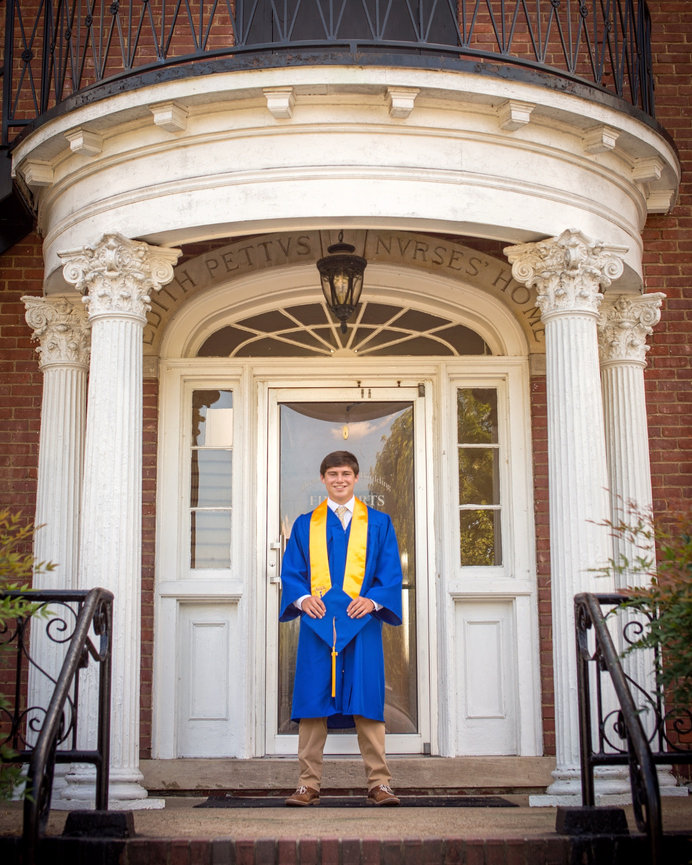 A graduate in a blue gown stands smiling in front of a historic building with white columns.