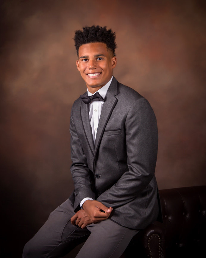 Young man smiling in a formal suit and bow tie, seated against a dark, blurred background.