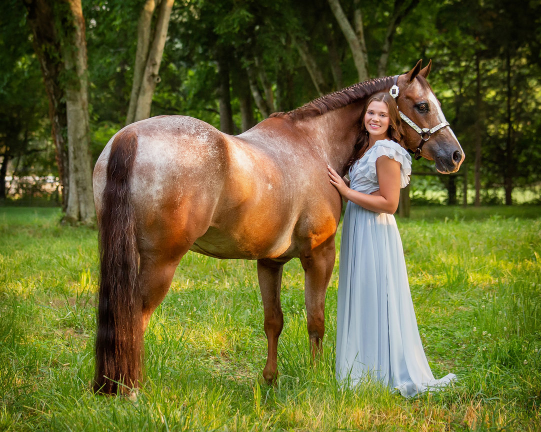 Young girl in a flowing dress stands beside a brown spotted horse in a lush green setting.