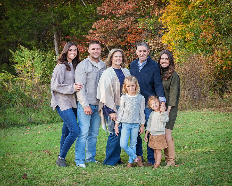 A family of seven posing together outdoors in a park with autumn foliage.
