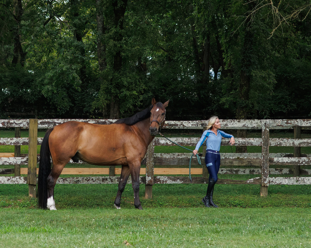 Woman leading a brown horse along a grassy area with a wooden fence and trees in the background.