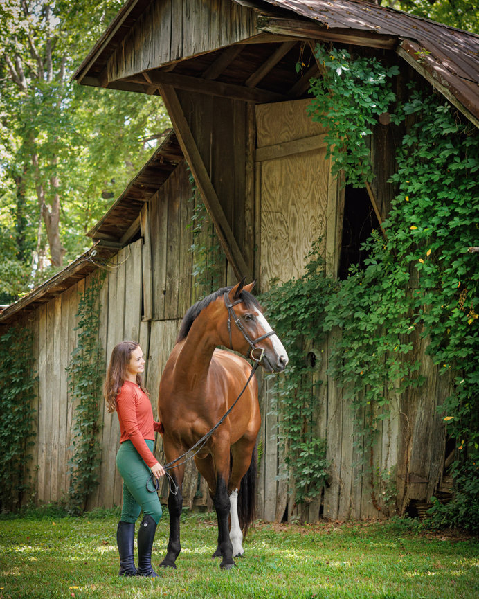A woman in riding attire stands beside a chestnut horse in front of a rustic barn covered in greenery.