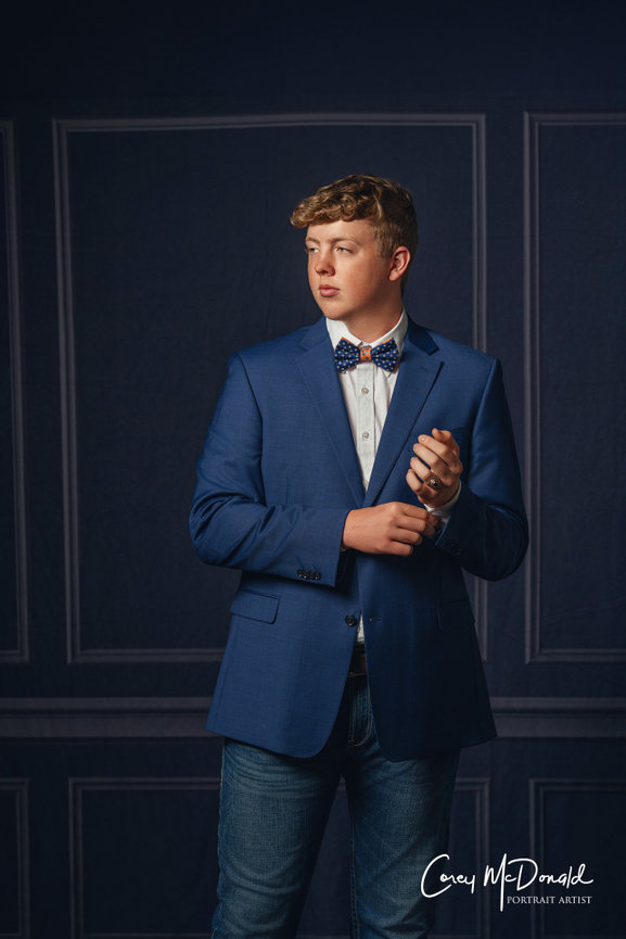 A young man in a blue suit with a bow tie stands against a dark, textured background.