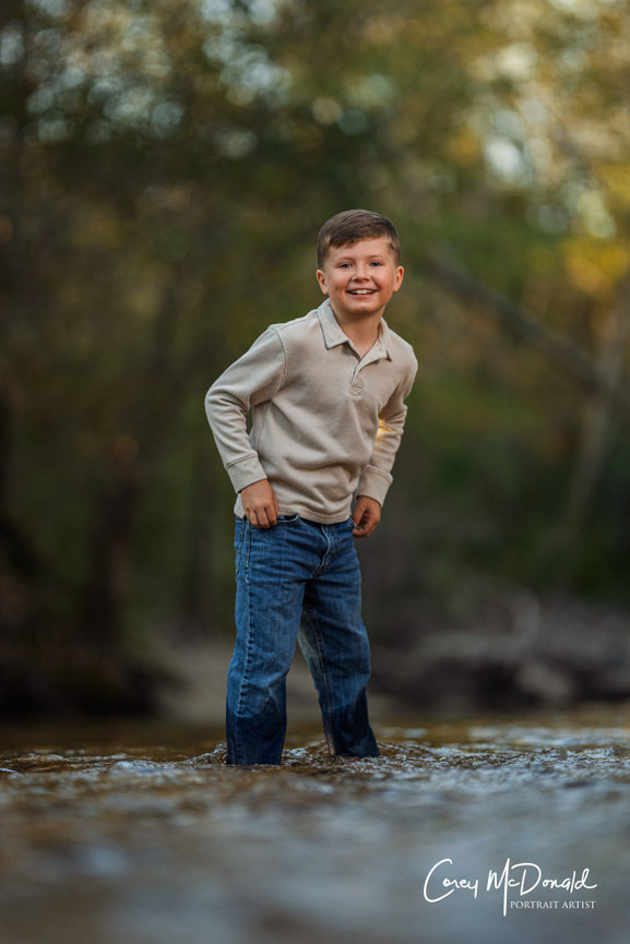 Boy smiling and standing in a shallow stream, wearing a gray shirt and jeans, with a blurred natural background.