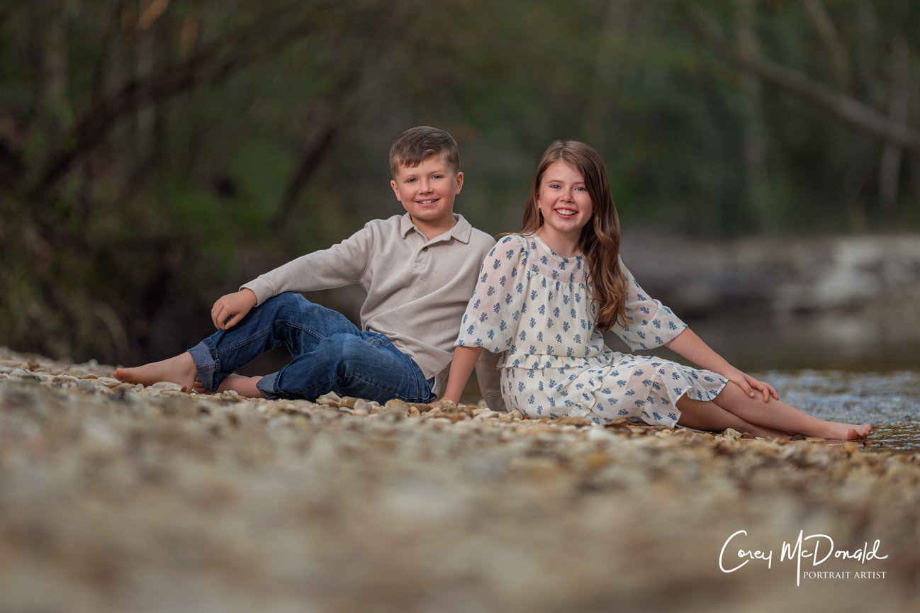Two children sitting on a rocky riverbank, smiling. The boy wears jeans and a beige shirt; the girl wears a patterned dress.