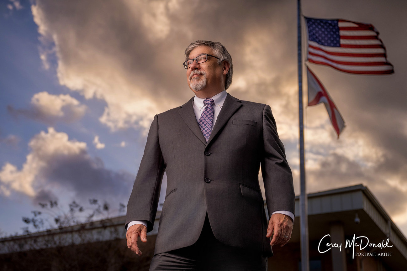 A man in a suit stands confidently outside with flags and a dramatic cloudy sky in the background.