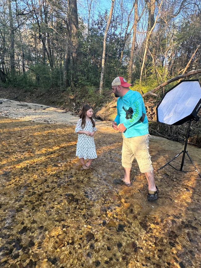 Man and child standing in a shallow creek with photo equipment; surrounded by trees in a natural setting.