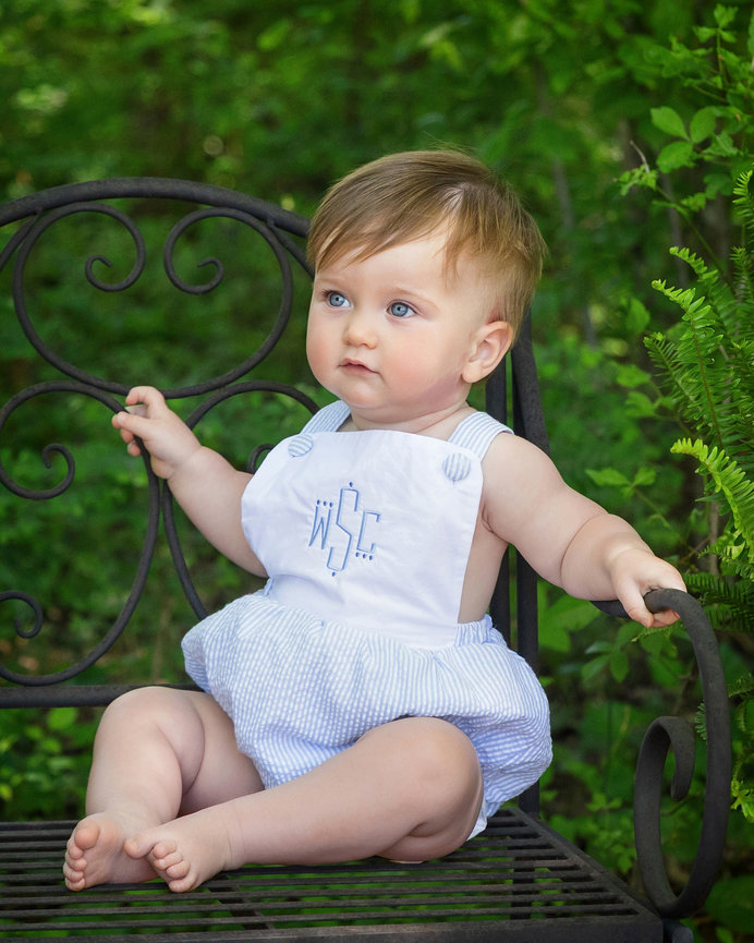 Baby sitting on a wrought iron bench in a garden, wearing a white romper with blue embroidery.
