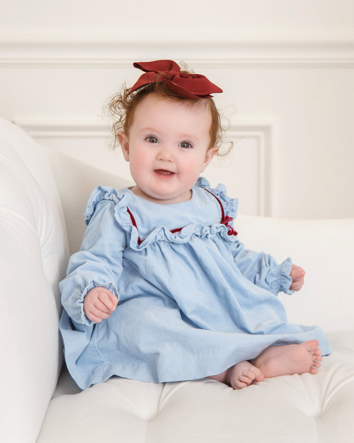 A baby in a blue dress with ruffles and a red bow headband sits on a white couch, smiling.