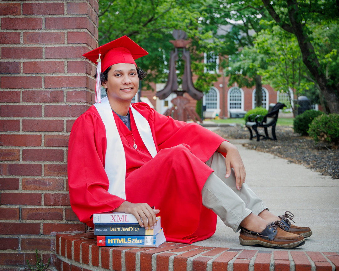 Young graduate in red cap and gown sits with books outside, near a brick wall and trees in the background.