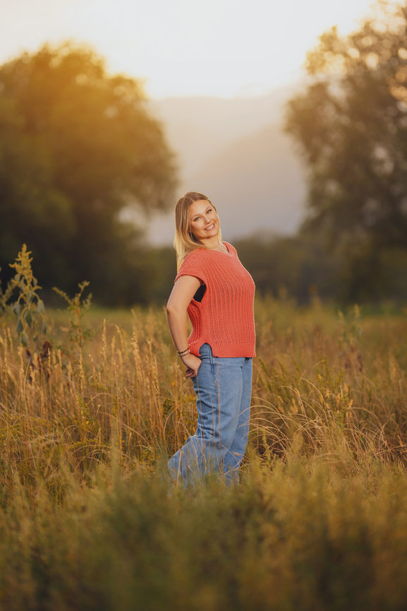Woman in orange top and jeans standing in a grassy field at sunset, smiling.