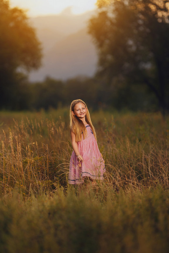 Young girl in a pink dress smiling in a sunlit field, surrounded by tall grasses and trees.