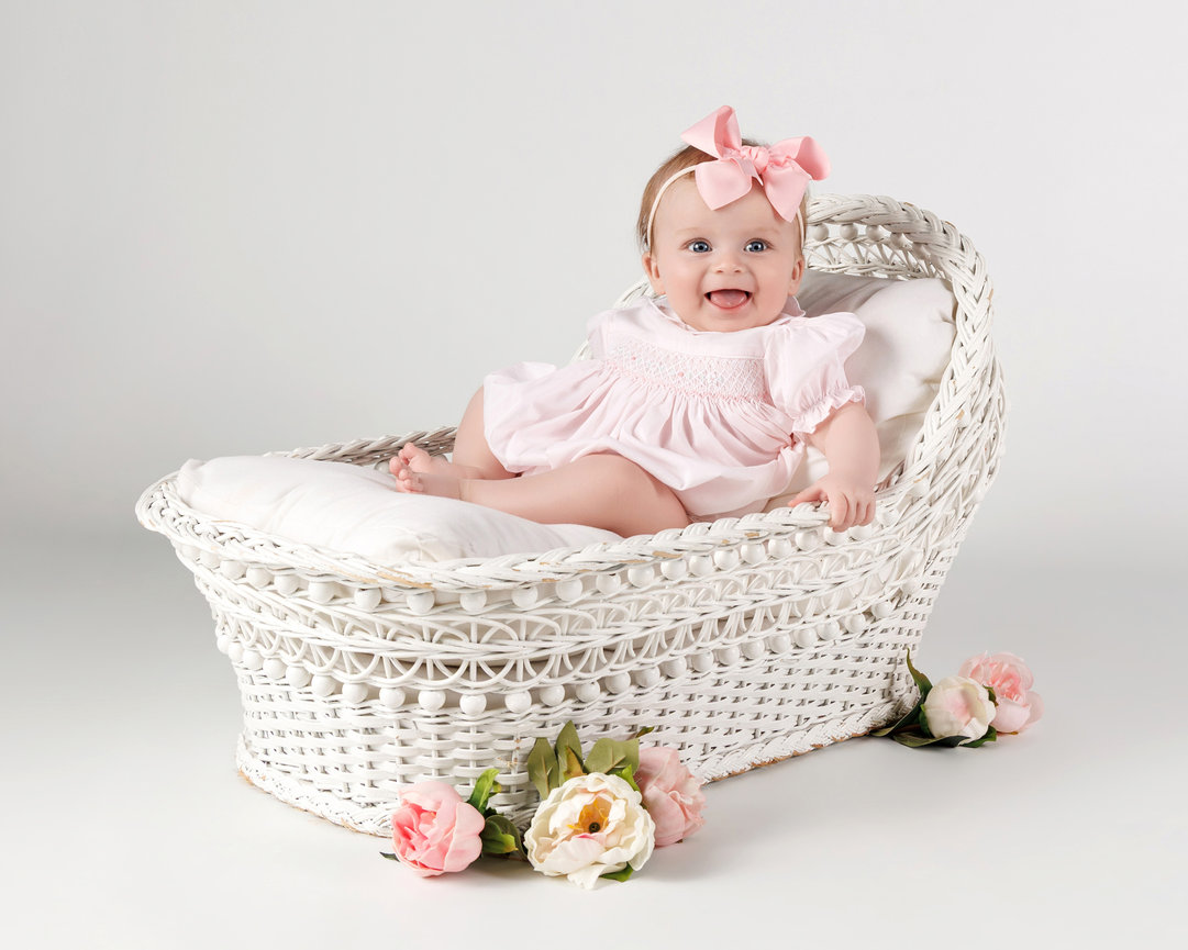 Smiling baby girl in pink dress and bow sitting in a white wicker basket with flowers