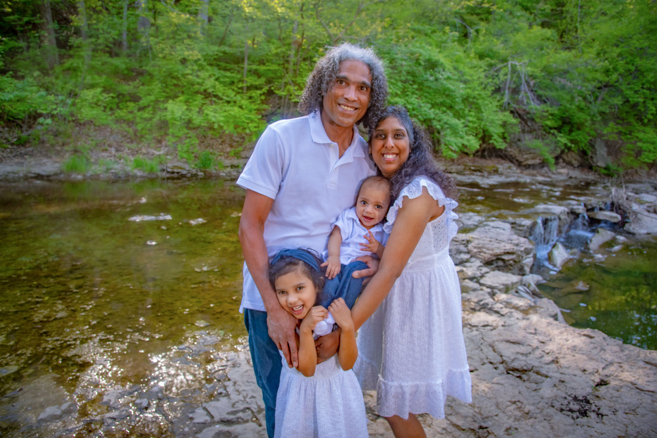 Family portrait by a river, featuring two adults and two young children, all smiling and wearing light-colored outfits.