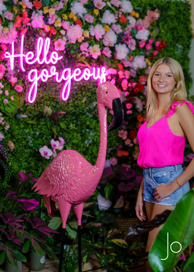 Young woman in pink tank top and denim shorts posing by a pink flamingo and neon Hello gorgeous sign, surrounded by plants.