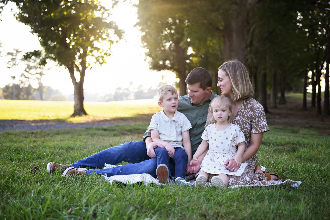 Family of four sitting on a blanket in an open field with trees and golden sunlight in the background during a relaxed outdoor session in Clemmons, NC at Tanglewood Park.
