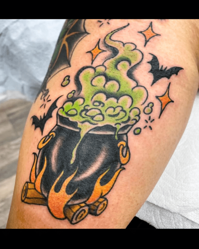 Color tattoo of a black cauldron over a log fire with green bubbles and smoke and rising from the cauldron and bats circling above.