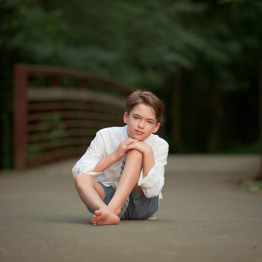 Young boy sitting outdoors in front of a rustic wooden bridge, captured in natural light with a timeless, fine art style.