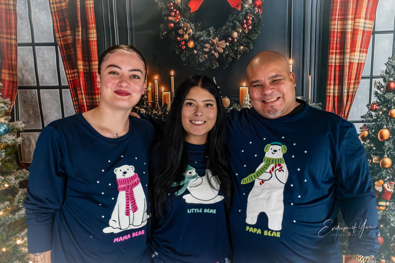Three people smile in matching holiday bear pajamas, standing in front of a festive backdrop with candles and a wreath.