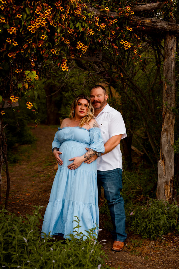 A couple poses in a garden, with the woman in a blue dress and the man in a white shirt, surrounded by lush greenery.