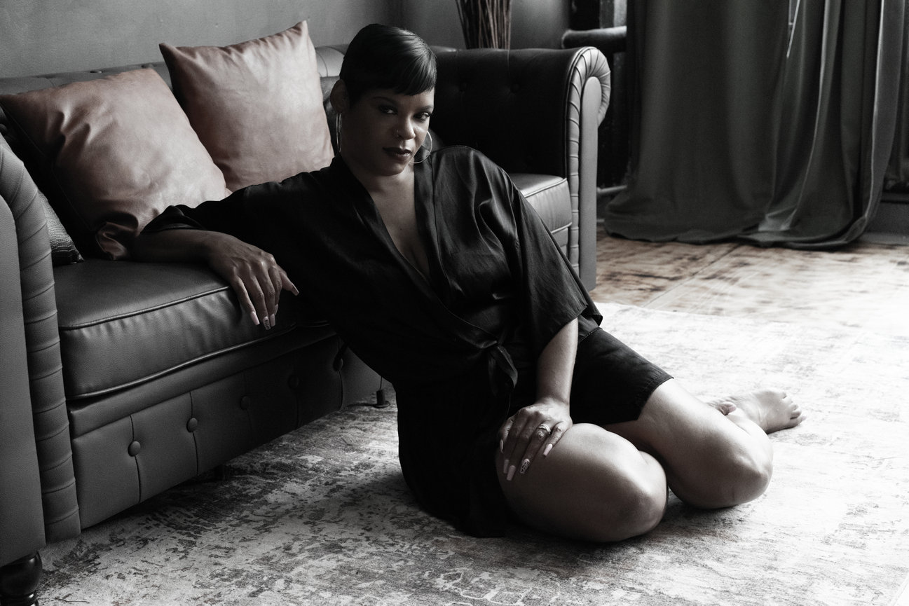 A woman in a black robe sits on the floor beside a sofa, posing elegantly in a stylish interior setting.