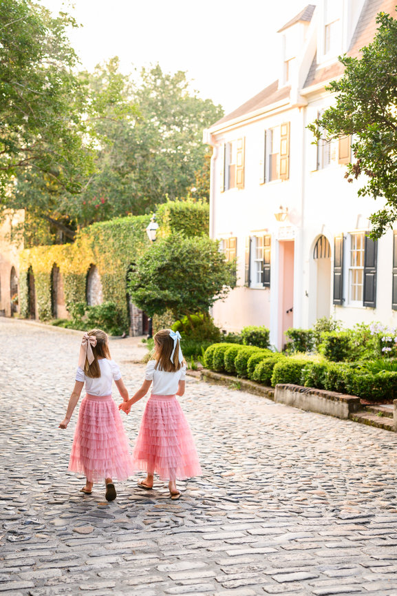 two little girls in matching pink skirts walk down a cobblestone street holding hands