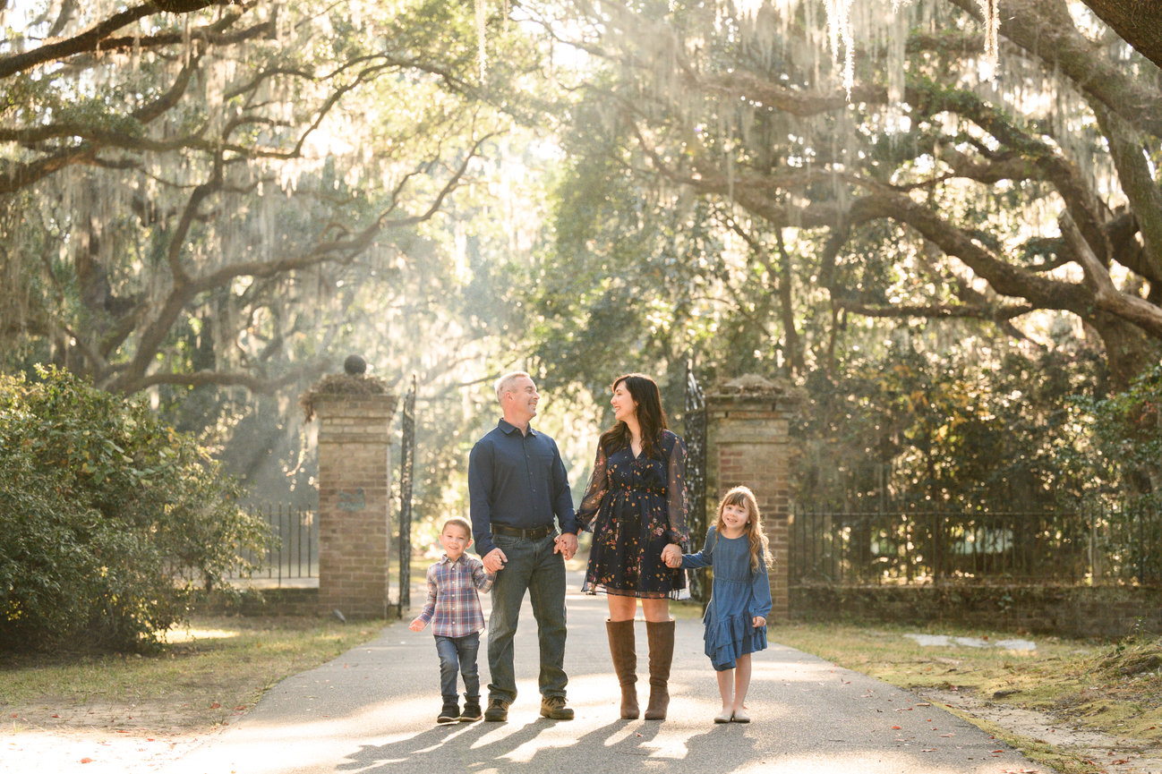 Family of four walking hand in hand on a sunlit path with trees and hanging moss in the background.