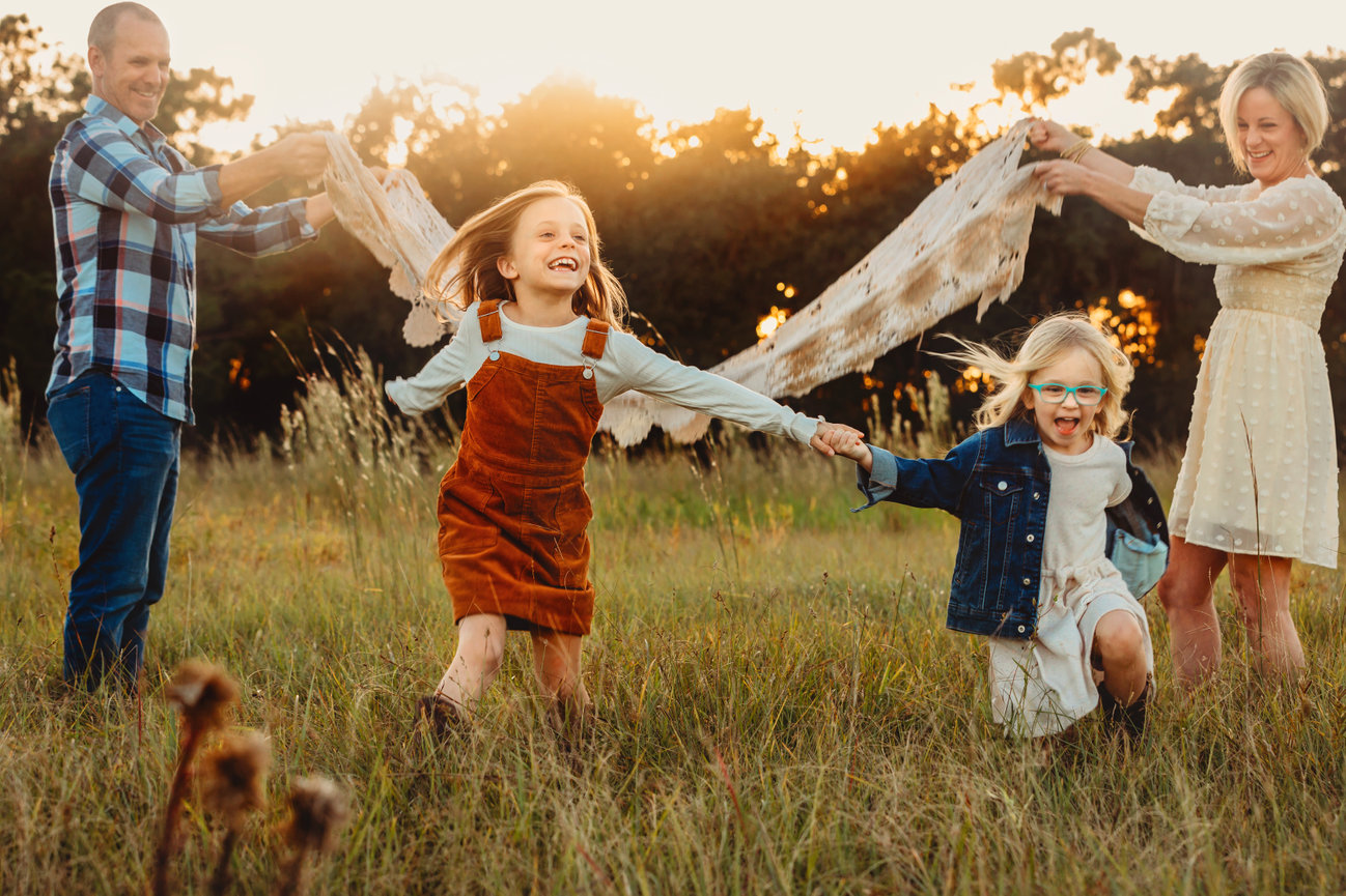 Family having fun in golden light while girls run under blanket in a field.