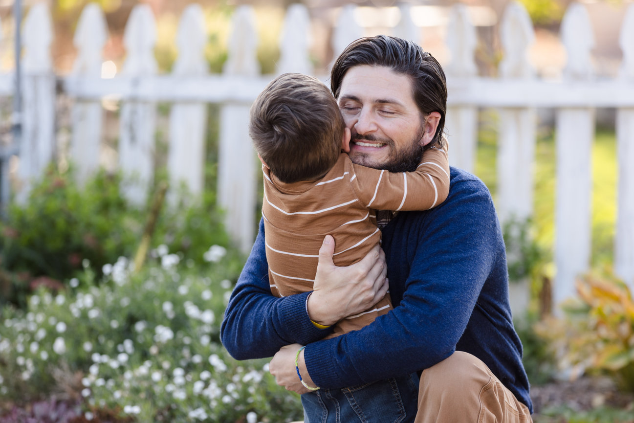 A dad and his toddler son are embracing as the dad kneels. There's an old picket fence behind them and flowers in the background.