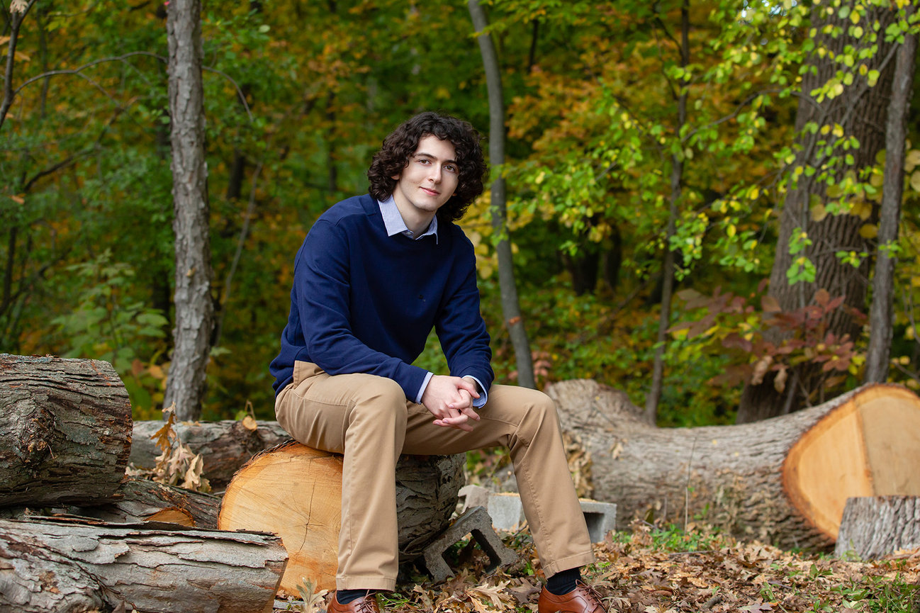 A high school senior boy is sitting on a freshly cut tree log in the woods.