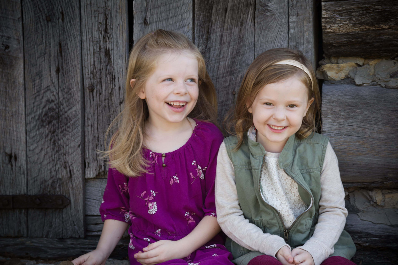Two smiling young girls sitting in front of a rustic wooden wall during a family photo session in Liberty photographed by Case Studios Photography