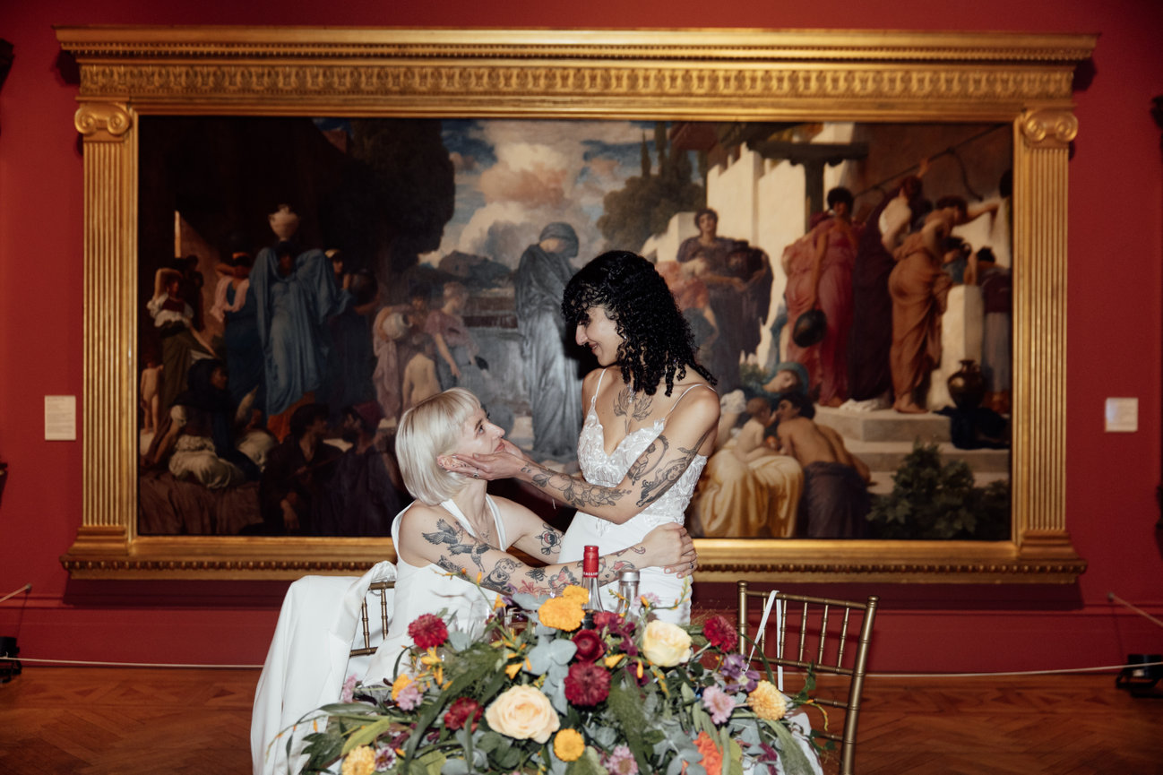 Direct flash editorial photography of two brides embrace at a floral-adorned table, positioned in front of a grand classical oil painting at Manchester Art Gallery.