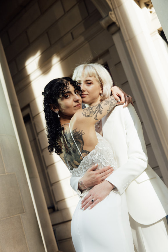 Two brides in modern wedding attire embracing in natural sunlight outside the neoclassical columns of Manchester Art Gallery.