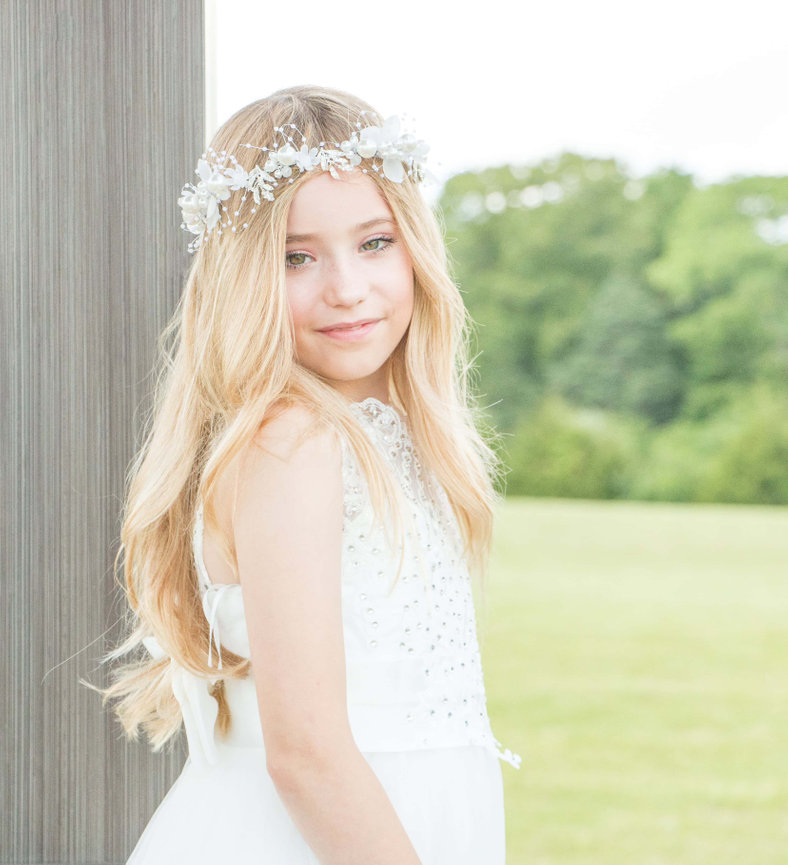 Young girl in a white dress with a flower crown, standing outdoors with a grassy background.