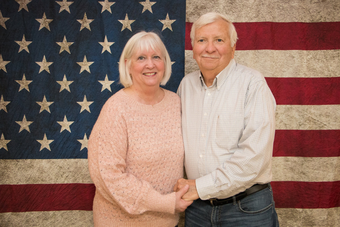 Older couple posing in front of a large American flag, holding hands and smiling warmly.