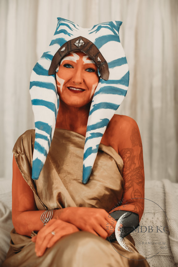 Smiling person in a blue and white striped headdress, wearing a tan shawl, seated indoors.
