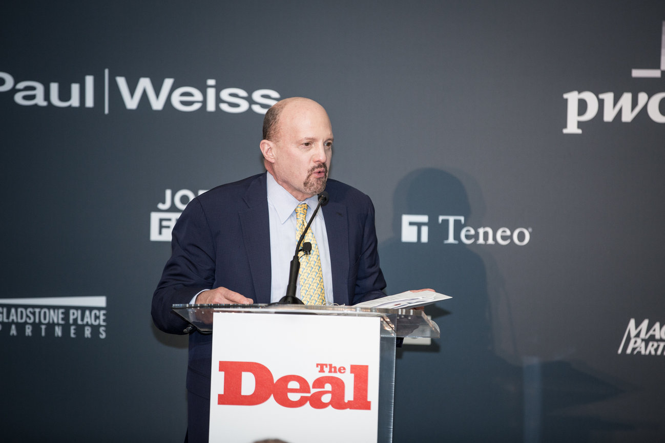 A man speaks at a podium with The Deal logo, set against a branded backdrop with various company names.