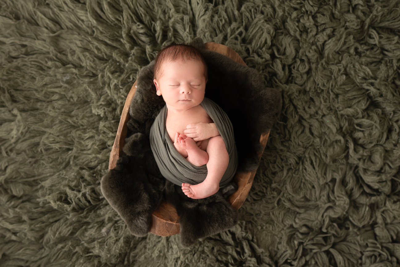 Baby wrapped in tactile knit fabric, nestled in rustic wood bowl with cozy layers during newborn session in Princeton, NJ