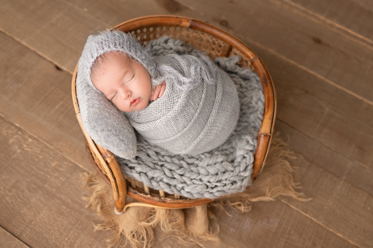 Fine art newborn photo of baby wrapped in cable-knit wrap and posed in papasan chair with soft layering