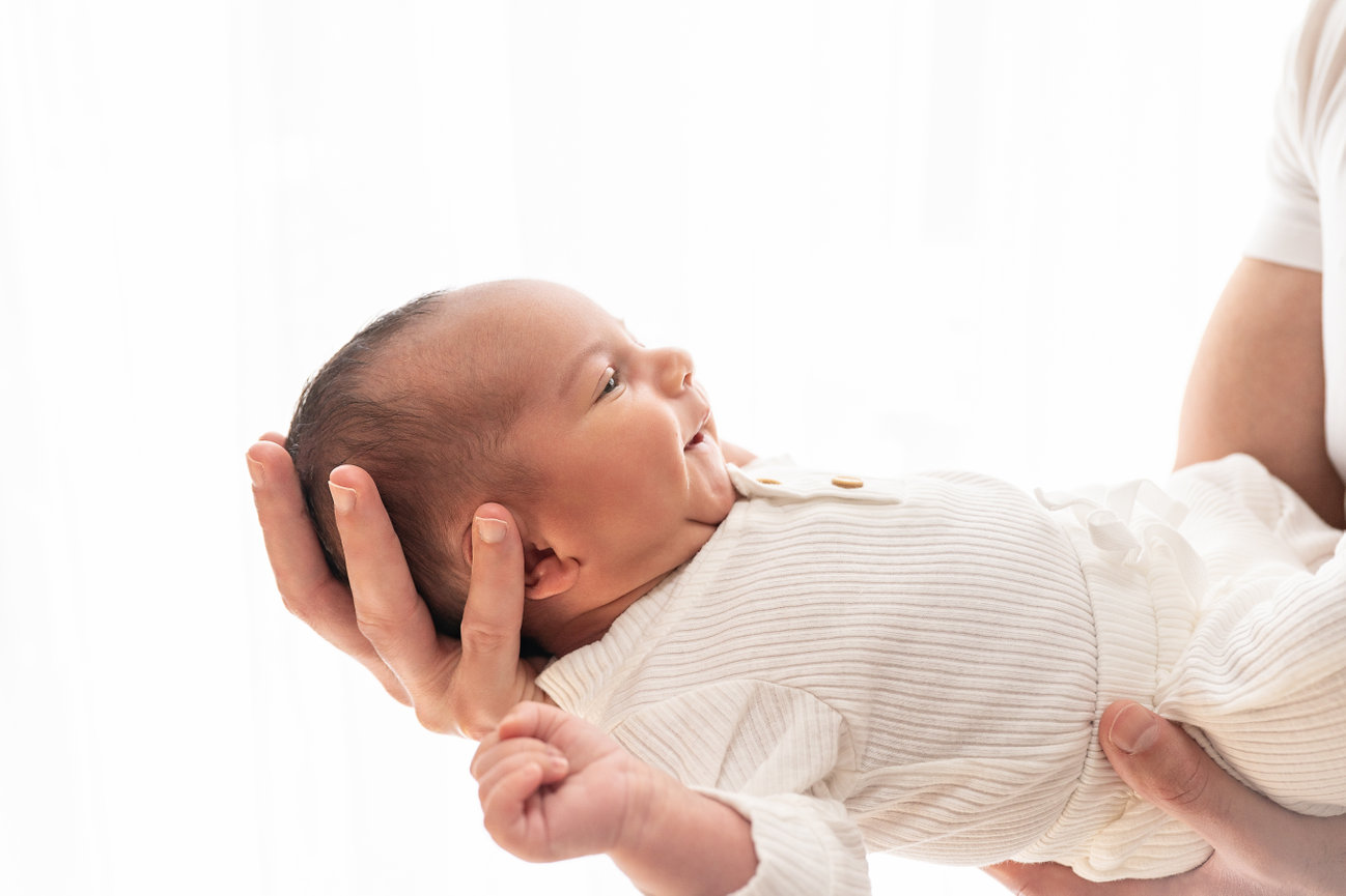 Parent holding newborn in white onesie near sheer window curtain