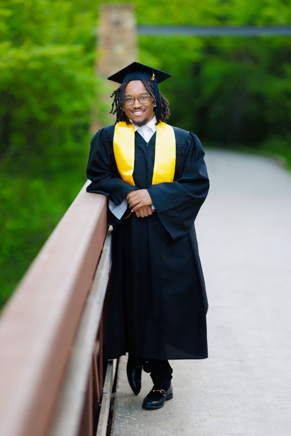 A graduate in a black cap and gown with a yellow stole stands on a bridge, smiling against a green backdrop at Oak Point Nature Preserve in Plano, Texas.
