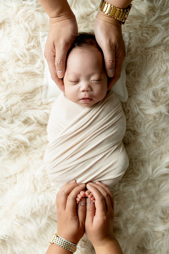Baby wrapped in a cream blanket, sleeping on a soft surface, with hands gently cradling the head and feet.
