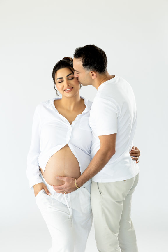 Pregnant woman in white, smiling, while a man kisses her forehead and touches her belly against a white background.